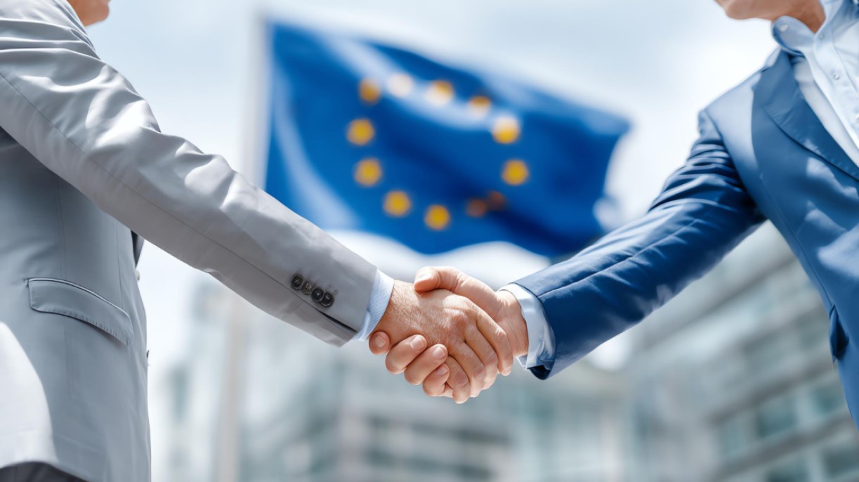 Two businesspeople shaking hands in front of the European Union flag, symbolizing partnership and agreement.