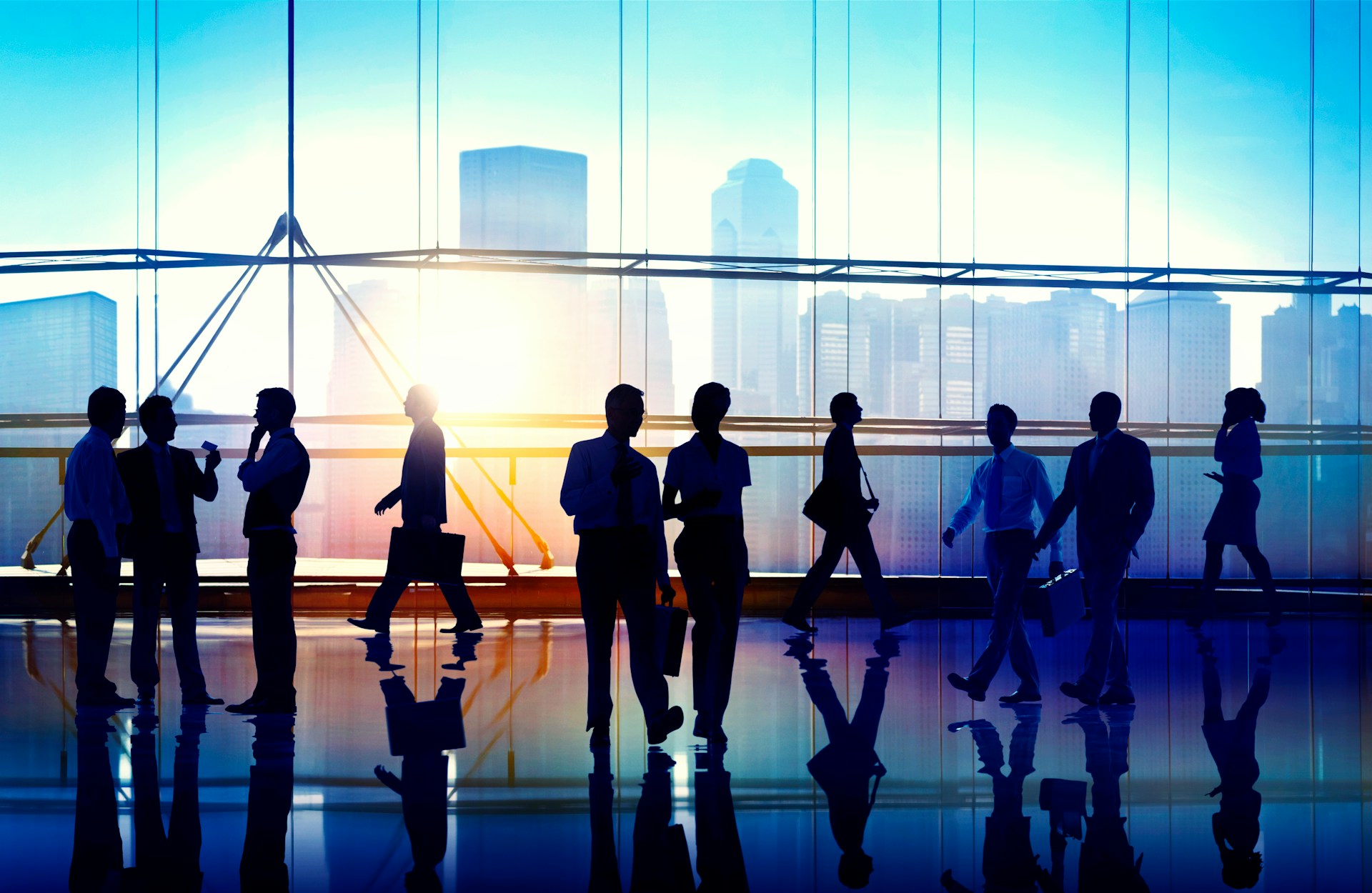 Silhouettes of businesspeople walking and talking inside a modern office with large windows showing a city skyline at sunrise.