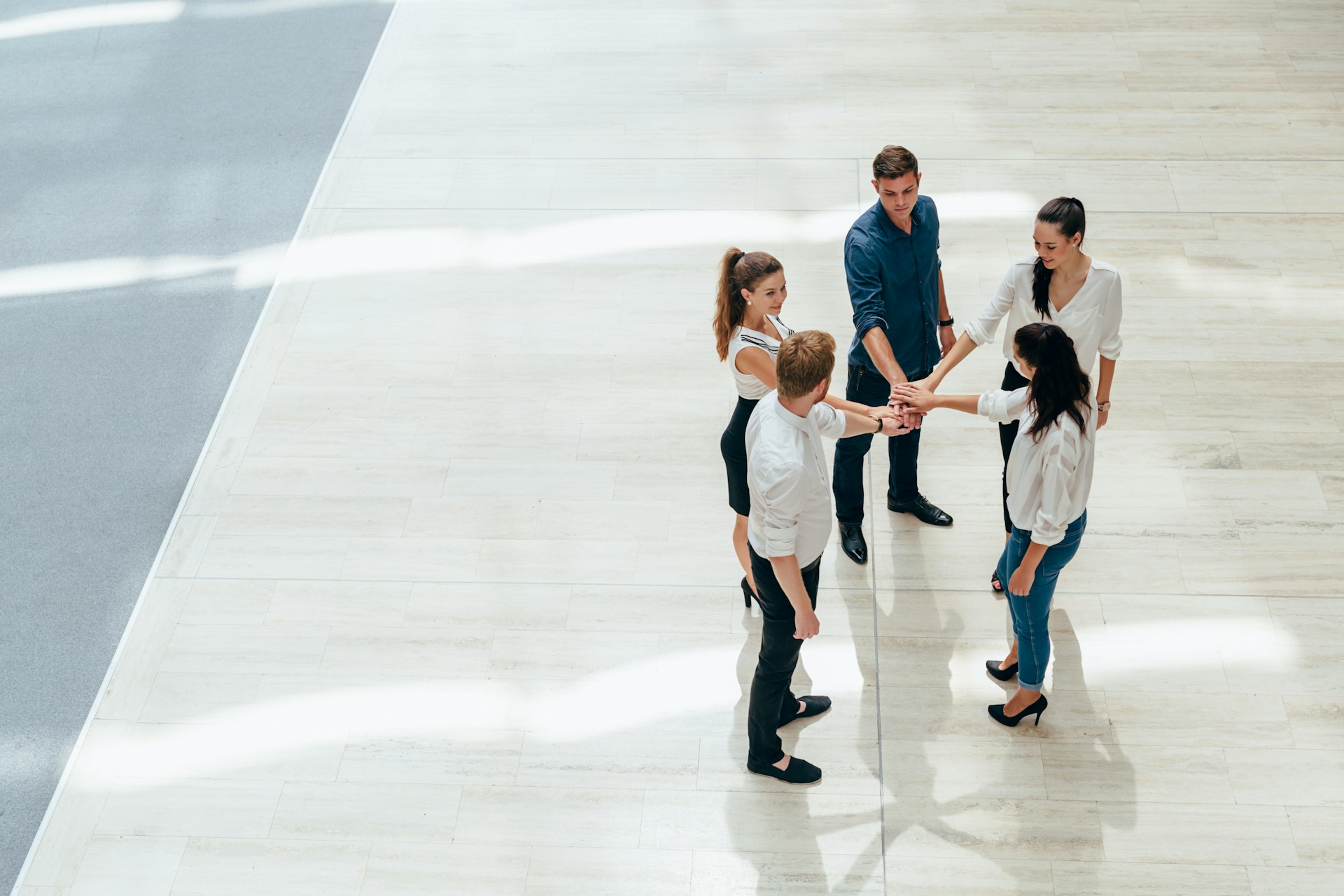 A group of five people standing in a circle, holding hands, in a well-lit indoor space with light-colored flooring.